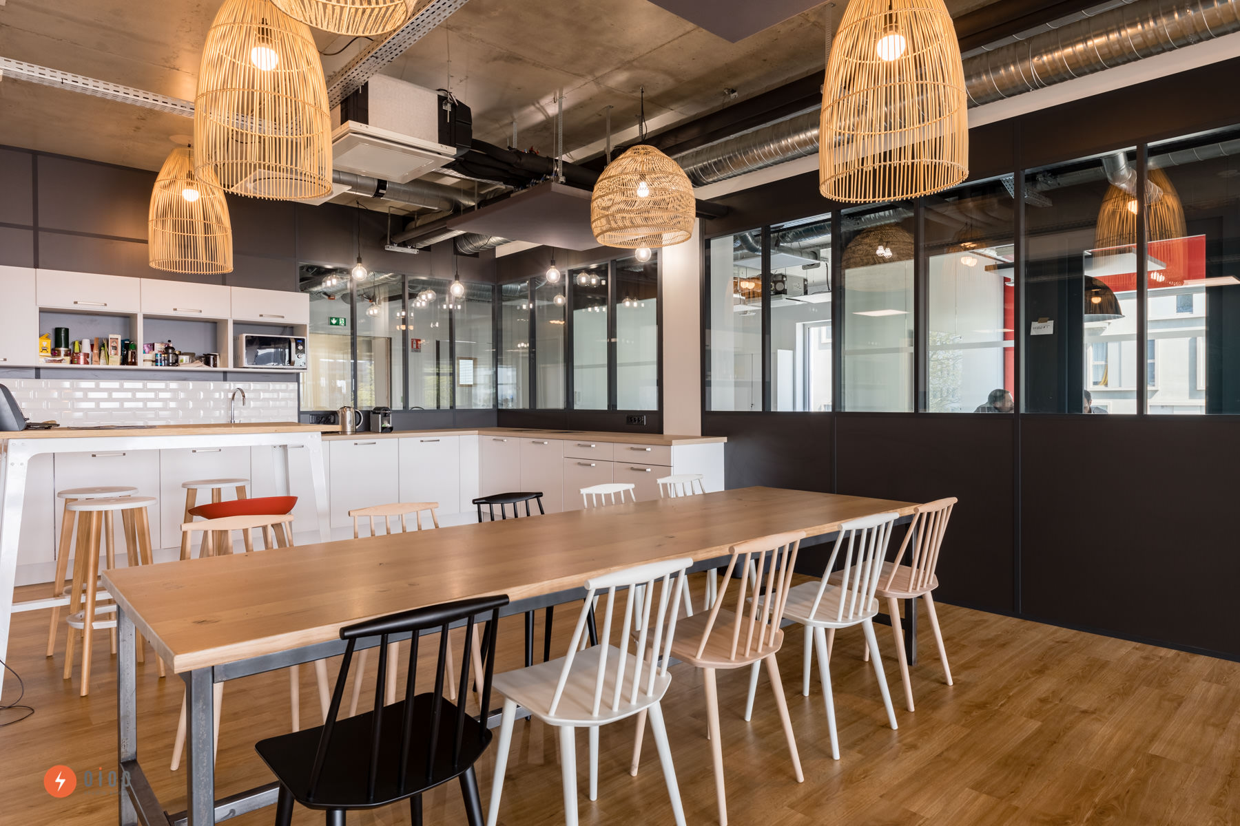 photo de design d'espace : vue d'un espace repas avec une grande table en bois au premier plan, une cuisine ouverte blanche en arrière plan, et une verrière noire qui sépare la cuisine de l'open space